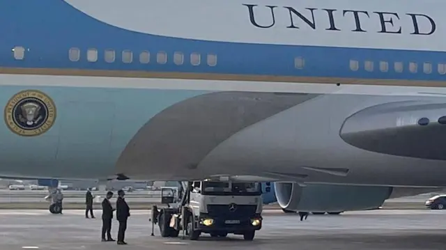 大統領印のついた専用機の下から、向こう側に横付けしているトラックが見える。周りにダークスーツ姿の職員が複数立っている。機体は白、青、水色に塗り分けられている