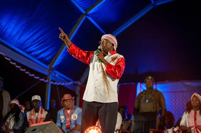Le président bissau-guinéen Umaru Sissoco Embalo en chemise blanche avec des manches longues rouges et un foulard autour de sa tête parle dans un micro dans sa main gauche et lève le doigt de sa main droit.