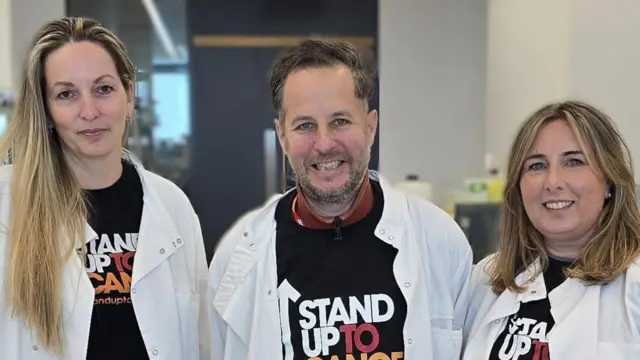 Зліва направо: проф. Петра Гамерлік, Алекс та Емма Девіс у лабораторії Манчестерського центру дослідження раку. Всі в білих халатах і усміхаються.