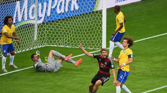 A imagem mostra um lance dentro da área durante uma partida entre Brasil e Alemanha. Enquanto um jogador alemão comemora de braços erguidos, jogadores brasileiros demonstram frustração ao redor da pequena área, com o goleiro caído no gramado segurando a bola e defensores observando a cena próximos à linha do gol.