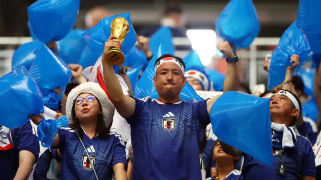 美国德州阿灵顿达拉斯球场FIFA世界杯F组日本对荷兰赛事上，一名男球迷（中）在举起充气蓝色塑料袋的球迷堆中举起双臂作势高呼，右手举起仿制的世界杯奖杯（14/6/2026）