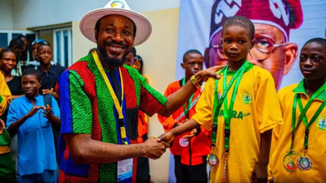 Yusuff Abdulsalam to wọ asọ alawọ ofeefe n bọ agbalagba ọkunrin kan lọwọ, ẹni to wọ aọ ankara alawọ eweko ati buluu.Awọn ọdọmọde to to mẹwa niye si lo rọgba yi wọn ka. Gbogbo wọn si lo gbe ami ẹyẹ sọrun gẹgẹ bii akopa ninu idije ere idaraya.