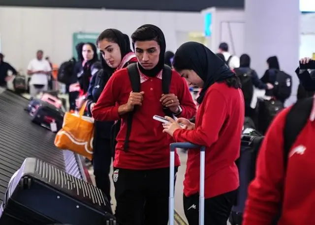 İranlı kadın sporcular kırmızı uzun kollu üst, siyah eşofman altı ve siyah yarım bağlanmış başörtüleriyle bagajlarını alıyor. 