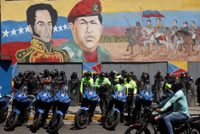 Fileiras de policiais de pé atrás de motos, usando capacetes em frente a um enorme mural pintado de Simón Bolívar e Hugo Chávez
