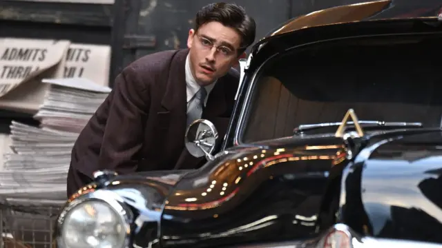 Timothée Chalamet, "Marty Supreme" filminin setinde, takım elbise ve gözlük takmış halde ve bir taksi penceresine doğru eğilmiş halde görülüyor.
