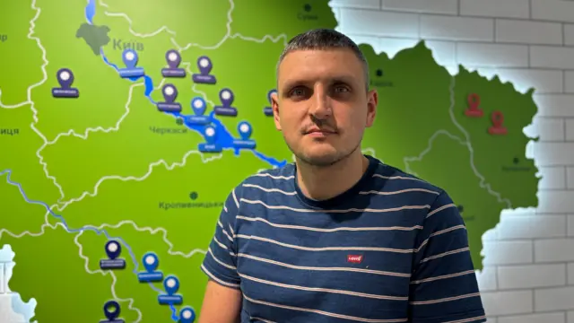 Чоловік у синьо-білій смугастій футболці стоїть перед картою України, яка світиться на стіні за його спиною.