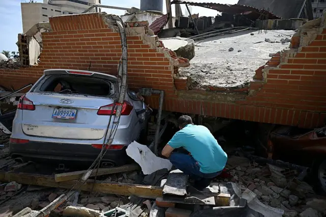 Un hombre acuclillado revisa el suelo alrededor de un auto aplastado por un pedazo de edificio que le cayó encima en los terremotos. 