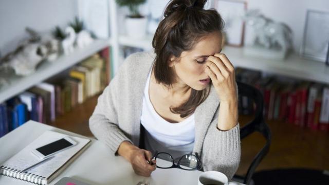 Una joven trabajadora desde su escritorio en casa sujeta sus gafas mientras que se lleva la mano izquierda a la cara en señal de cansancio.