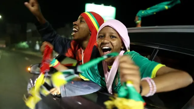 Deux supporters sénégalais vêtus des couleurs nationales applaudissent depuis une voiture en mouvement.