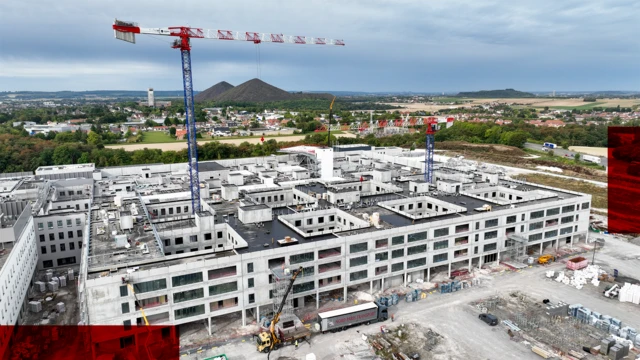 Аерофотознімок великої лікарні, що будується в Лоосі, поблизу Арраса. Будівля світло-сірого кольору, на території знаходяться будівельні машини та два великі крани. На задньому плані зображення видно сільську місцевість та кілька невисоких пагорбів.