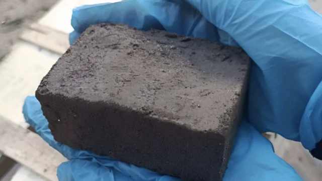 A close-up shot of the large, dark brown brick held by a pair of hands in blue surgical gloves.