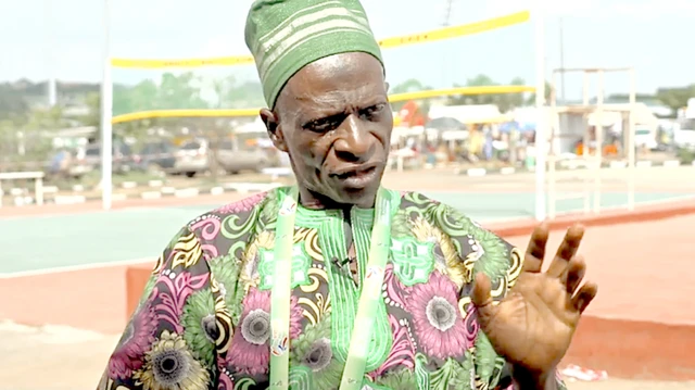Photo d'un homme d'une cinquantaine d'années, vêtu d'un tissu africain à fleurs largement roses et vertes et d'un chapeau vert, lève légèrement la main gauche pour montrer sa paume à la caméra lors d'un entretien. Un court d'Abula est visible à l'arrière-plan, avec un filet à garniture jaune en haut et en bas.