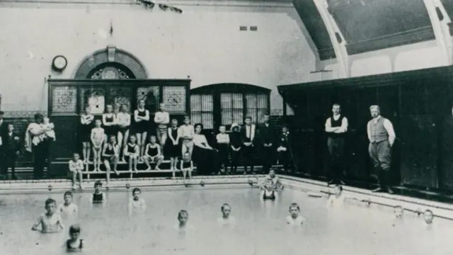 Una fotografía antigua en blanco y negro del interior de los baños. Se ven varios niños en traje de baño sentados en bancos junto a la piscina, muchos de ellos también en el agua.