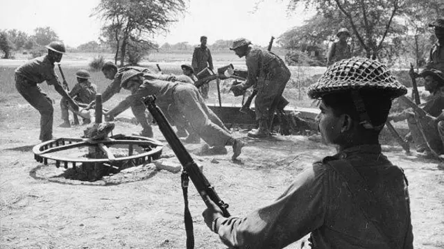 বন্দুক হাতে যুদ্ধের প্রস্তুতি নিচ্ছেন কয়েকজন সেনা, অন্য কয়েকজন চাকার মতো দেখতে একটি জিনিস নিয়ে কাজ করছেন 