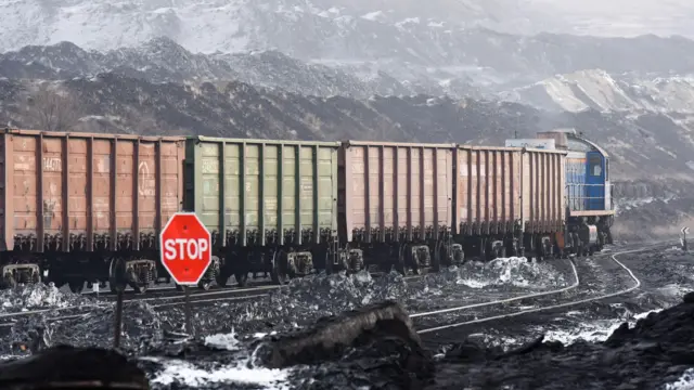 На заднем плане - возвышенности, на которых кое-где лежит снег. На среднем плане - грузовой поезд везет вагоны красного и зеленого цветов с слева-направо. На переднем плане красный знак с белой надписью Stop.