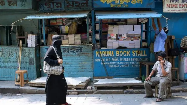 একটি বইয়ের দোকানের সামনে দিয়ে হেঁটে যাচ্ছেন বোরকা পরা একজন নারী। পাশে একটি চেয়ারের বসে ও তার পাশে দাঁড়িয়ে আছেন দুই জন পুরুষ।