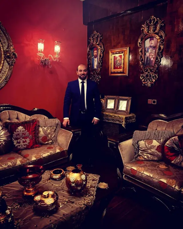 II. Abdülhamid'in torunu İbrahim Jour, evindeki kırmızı duvarlı bir odada Osmanlı aile yadigarlarının önünde takım elbiseyle poz veriyor.
