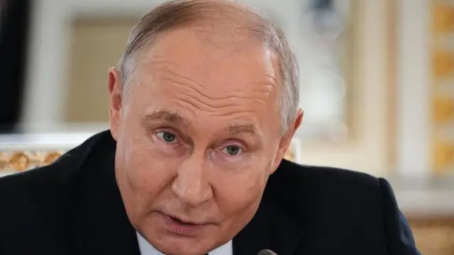 Putin ubrany w ciemnoniebieski garnitur, jasnoniebieską koszulę i krawat w kropki pochyla się nad mikrofonem na konferencji prasowej, unosząc brwi