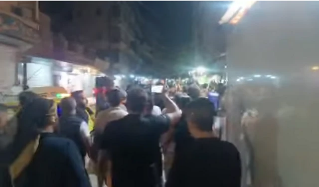 Bulanık ekran görüntüsünde yürüyüş yapan grup arkadan görünüyor, akşam ve sokak lambası yanıyor.