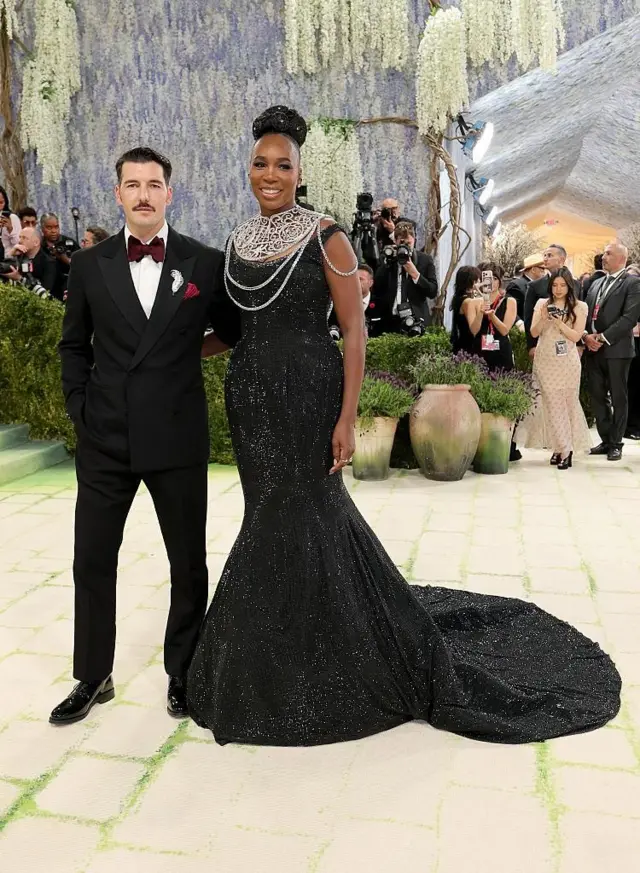 Un hombre y una mujer posan en una alfombra verde durante un evento formal; él viste traje negro con pajarita y ella un vestido largo negro con joyería. Hay fotógrafos y decoración floral de fondo.