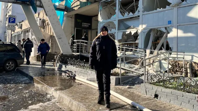 Жінка в синій куртці та капелюсі стоїть перед пошкодженою будівлею