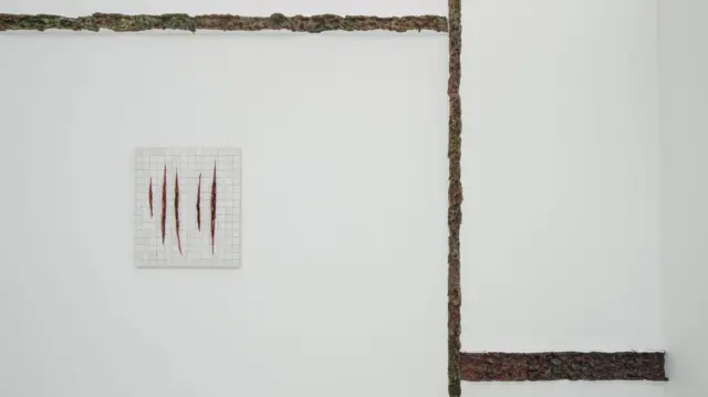Em uma parede branca minimalista, uma pequena obra de azulejos exibe cortes verticais que lembram feridas abertas, enquanto estruturas orgânicas e escuras percorrem o espaço como fissuras ou cicatrizes.