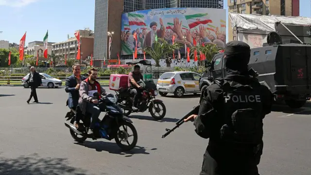 یکی از میدانهای تهران در حالیکه مامور پلیسی که صورت خود را پوشانده و اسلحه در دست دارد ایستاده. عابران و دو موتور سوار در حال گذار از خیابان هستند.