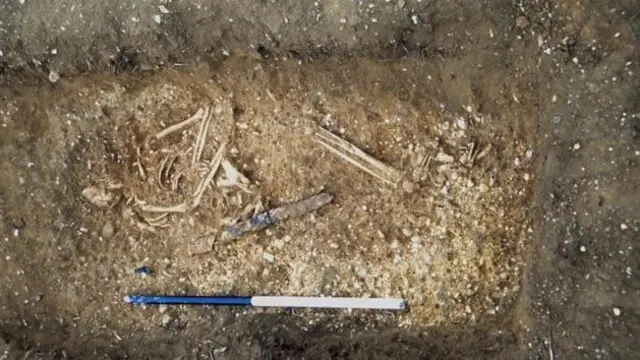 Excavación del esqueleto de un hombre que yace de costado, con la cabeza hacia la izquierda y las piernas flexionadas. Debajo se encuentran los restos de una espada. Se ha colocado un poste azul y blanco debajo de los restos.