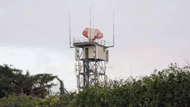 En primer plano se observa una torre metálica que se alza sobre un arbusto. La torre tiene una antena parabólica roja y blanca en la parte superior.