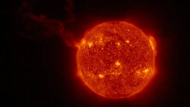 Güneş, yüzeyinde parlak sarı noktalar bulunan kırmızı bir top olarak görülüyor; soldan uzun bir bulut görünüyor.