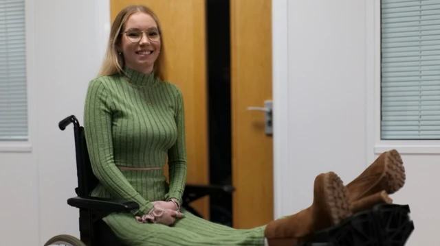 Megan está sentada em uma cadeira de rodas, olhando para a câmera e sorrindo. As pernas dela estão elevadas e apoiadas em uma estrutura. Ela usa óculos de grau, roupa verde e botas. 