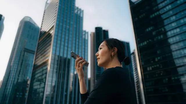 Azjatycka bizneswoman rozmawia z wirtualnym asystentem na swoim smartfonie. Jest na zewnątrz, a za nią widać liczne wieżowce.