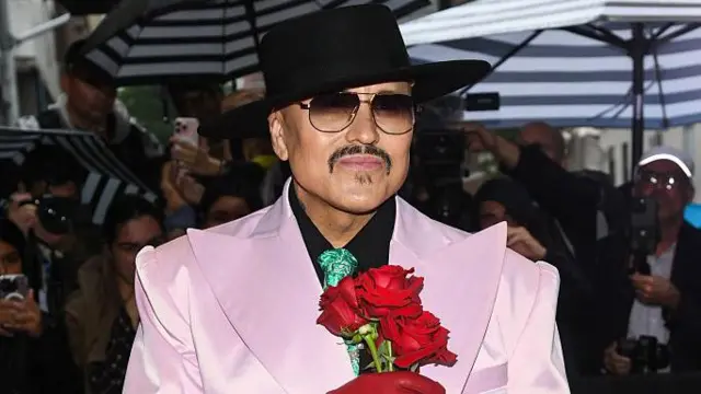 Willy Chavarria no tapete vermelho, com chapéu preto, óculos escuros e smoking rosa-claro com lapelas largas, segurando rosas vermelhas, cercado por fotógrafos