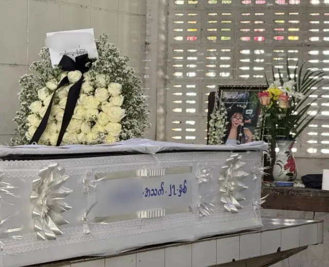 မေ့ဆေးလွန်ပြီး သေဆုံးခဲ့တဲ့ မနေခြည်ရဲ့ ဈာပနမြင်ကွင်း၊ အဖြူရောင် ရုပ်အလောင်းထည့် အခေါင်း တစ်ခုမှာ အသက် ၂၇ နှစ်လို့ ရေးထိုးထားပါတယ်။ ရုပ်အလောင်းရဲ့ အပေါ် ဘက်မှာတော့ မျက်မှန်တပ်ထားတဲ့ အမျိုးသမီးငယ်တစ်ယောက်ရဲ့ ဓာတ်ပုံကို ထောင်ထားပြီး အဖြူရောင်နှင်းဆီတွေနဲ့ လုပ်ထားတဲ့ ပန်းခွေတစ်ခုကို အခေါင်း ပေါ်မှာ တင်ထားပါတယ်။