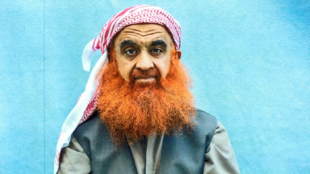 Фотографія Халіда Шейха Мохаммеда. У нього довга руда борода, біло-червона хустка на голові, сіра сорочка та темно-сіра безрукавка