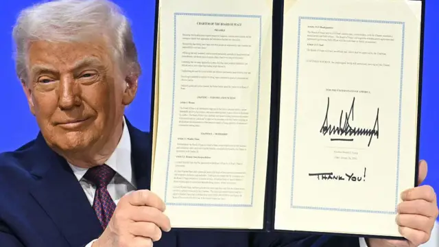 US President Donald Trump holds a large printed statement with his signature at the bottom during the World Economic Forum annual meeting in Davos on 22 January 2026. He is smiling and holding the two-page statement bound in a display book with both hands.