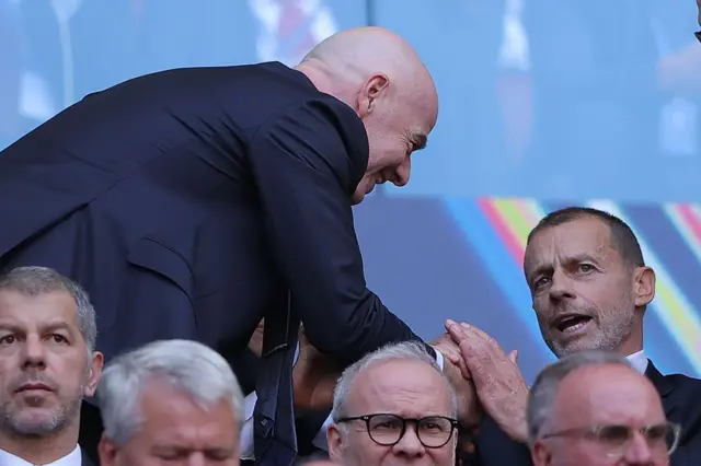 FIFA Başkanı Gianni Infantino ve UEFA Başkanı Aleksander Ceferin, tribünlerde el sıkışarak konuşuyorlar.
