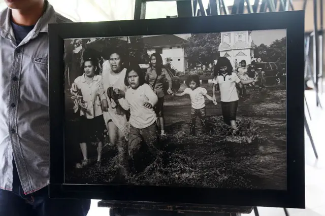 Fotografia enquadrada mostra crianças atravessando área alagada com mulheres. 