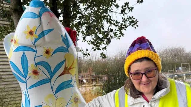 Una mujer que lleva un gorro de colores brillantes, gafas y un chaleco reflectante está de pie junto a la escultura de una rata florida.