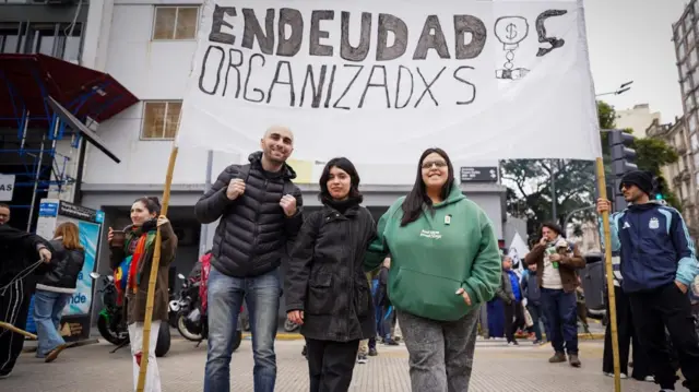 Tres personas frente a un cartel que dice Endeudados Oranizados.