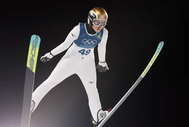 白いウェアに「49」のゼッケンをつけた丸山が両足にスキーをつけ、両手両足を開き、暗い空を背に飛んでいる