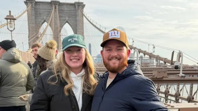 Egy nő és egy férfi áll egy New York-i hídon, mosolyogva a kamerába. Mindketten kabátot és baseballsapkát viselnek.