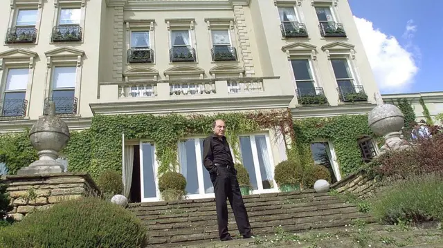 
Mwanamume aliyevaa nguo nyeusi anasimama kwenye ngazi mbele ya nyumba nzuri