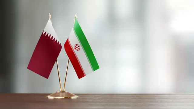 علما قطر وإيران بحجم صغير على قاعدة ذهبية دائرية بحجم صغير موضوعة على مكتب، لونه من درجات البني الفاتح مع خلفية ضبابية. 