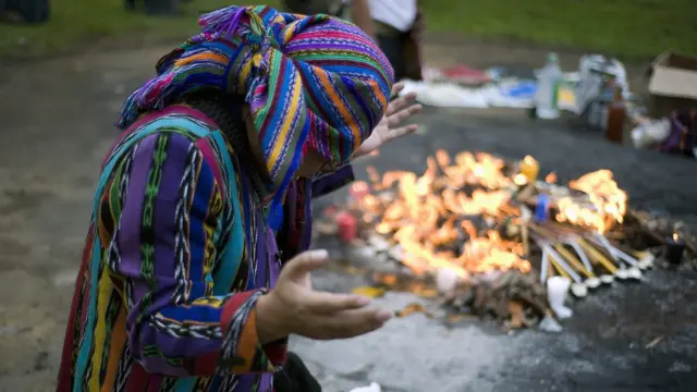 Una persona vestida con atuendos tradicionales mayas realiza una ceremonia al lado de una fogata