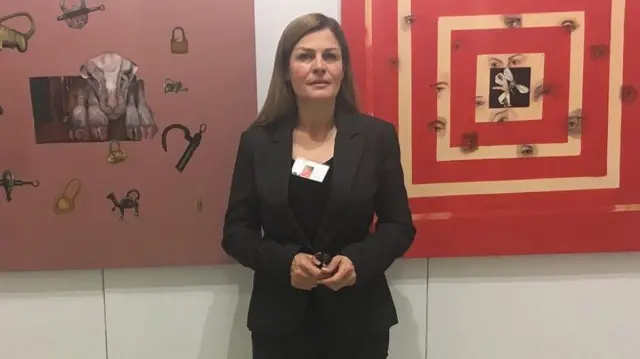 مهرانگیز ایمن‌پور لباس مشکی به تن دارد و برگه شناسایی بازدیدکننده به پیراهنش سنجاق شده است. او مقابل دیواری به رنگ بژ ایستاده که دو اثر هنری قرمز رنگ بر آن آویزان است.