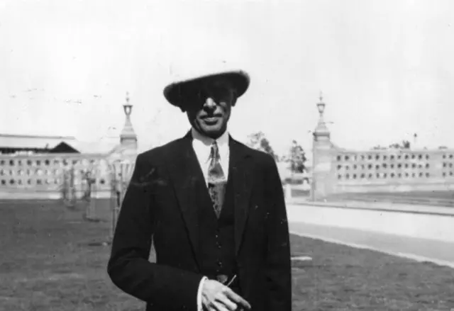 মধ্যবয়সী মুহাম্মদ আলী জিন্নাহ কোট ও মাথায় টুপি পরে একটি মাঠে দাঁড়িয়ে আছেন, পেছনে দূরে একটি দেয়াল দেখা যাচ্ছে। তার একটি হাত পেটের কাছে ভাঁজ করা ও মুখে হালকা হাসি