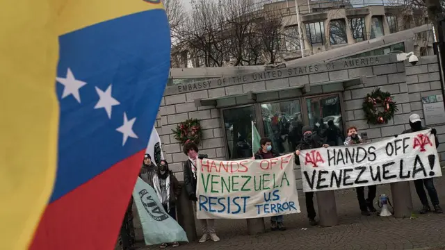 Bandeira venezuelana em primeiro plano, com pessoas ao fundo se manifestando com cartazes com os dizeres "afaste suas mãos da Venezuela", em inglês