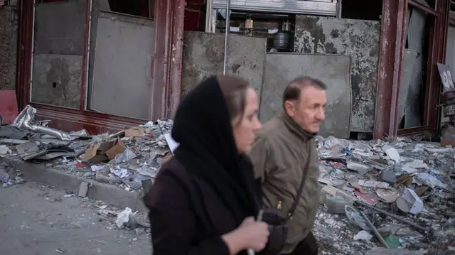 एक बुज़ुर्ग ईरानी व्यक्ति और एक महिला 2 मार्च 2026 को ईरान के तेहरान में अमेरिकी-इज़राइली हमलों में पूरी तरह तबाह हो चुकी पुलिस मुख्यालय के पास स्थित एक ध्वस्त इमारत के सामने से गुजरते हुए