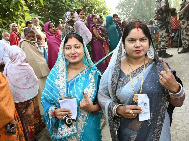 শাড়ি ও মাথায় সিঁদুর পরা দুইজন নারী একহাতে পরিচয়পত্র ধরে অন্য হাতের আঙুল তুলে ভোট দেওয়ার চিহ্ন অমোচনীয় কালি দেখাচ্ছেন। পেছনে নারীদের লাইন। লাইনের পেছনে কয়েকজন পুরুষ এবং অন্য পাশে আইনশৃঙ্খলা বাহিনীর দুই জন সদস্য দাঁড়িয়ে আছেন।